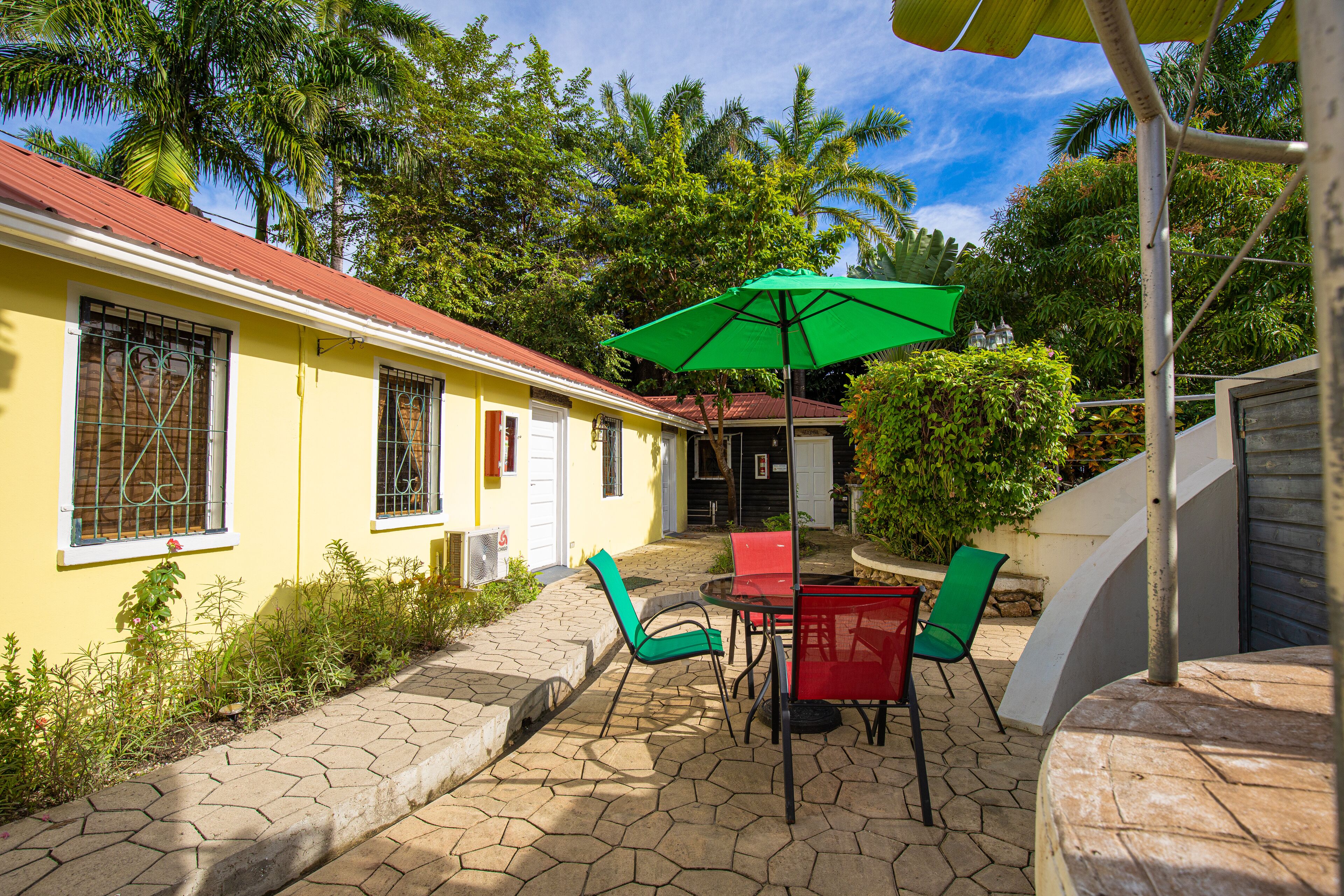 A yellow building with a red roof, a patio with a table and chairs, and a green umbrella.