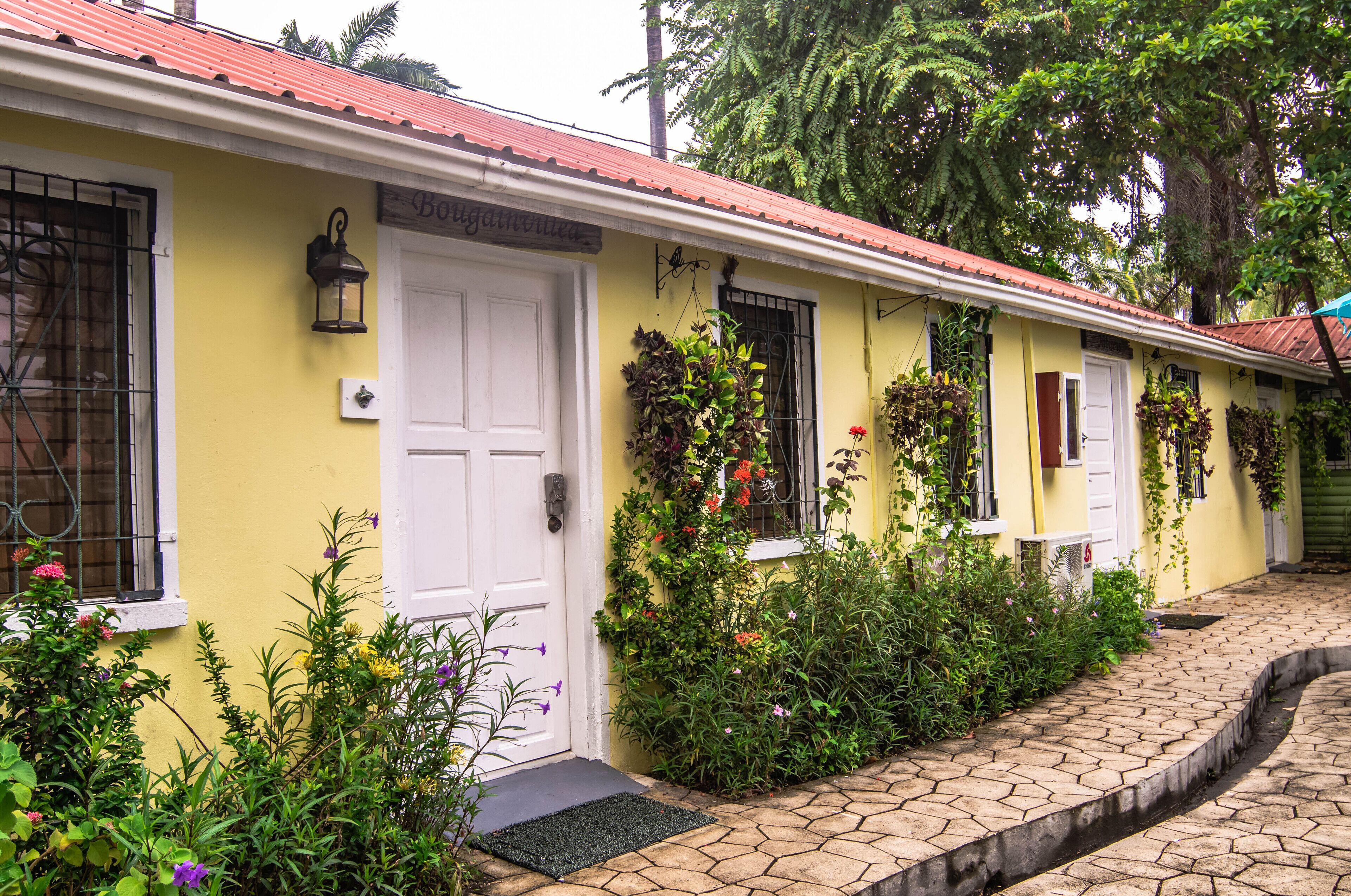 A yellow building with a red roof, white doors, and windows with metal bars. There is a small garden with various plants and flowers in front of the building.