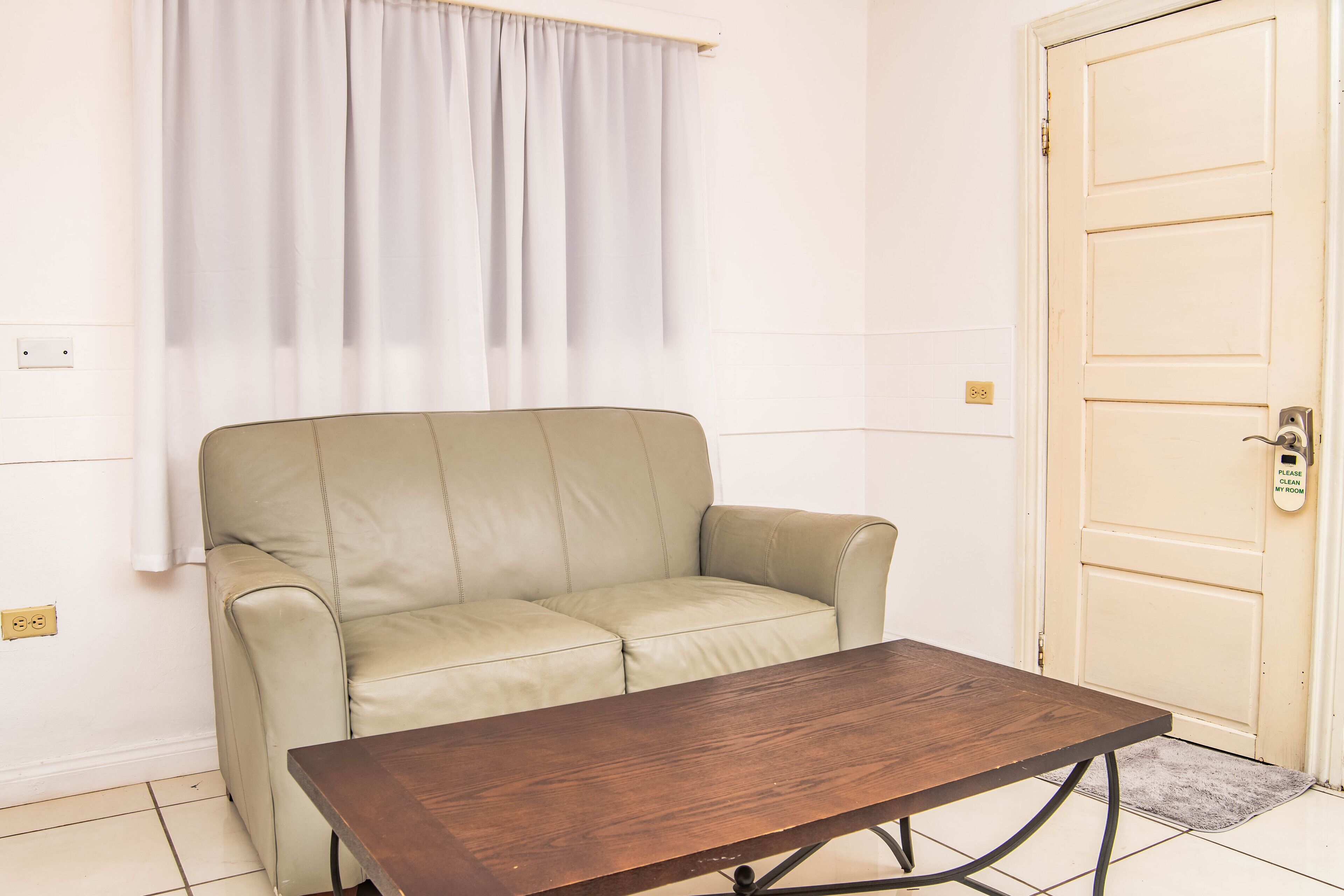 A beige leather sofa, a wooden table with metal legs, a white door with a "Please clean my room" sign, and a white curtain.
