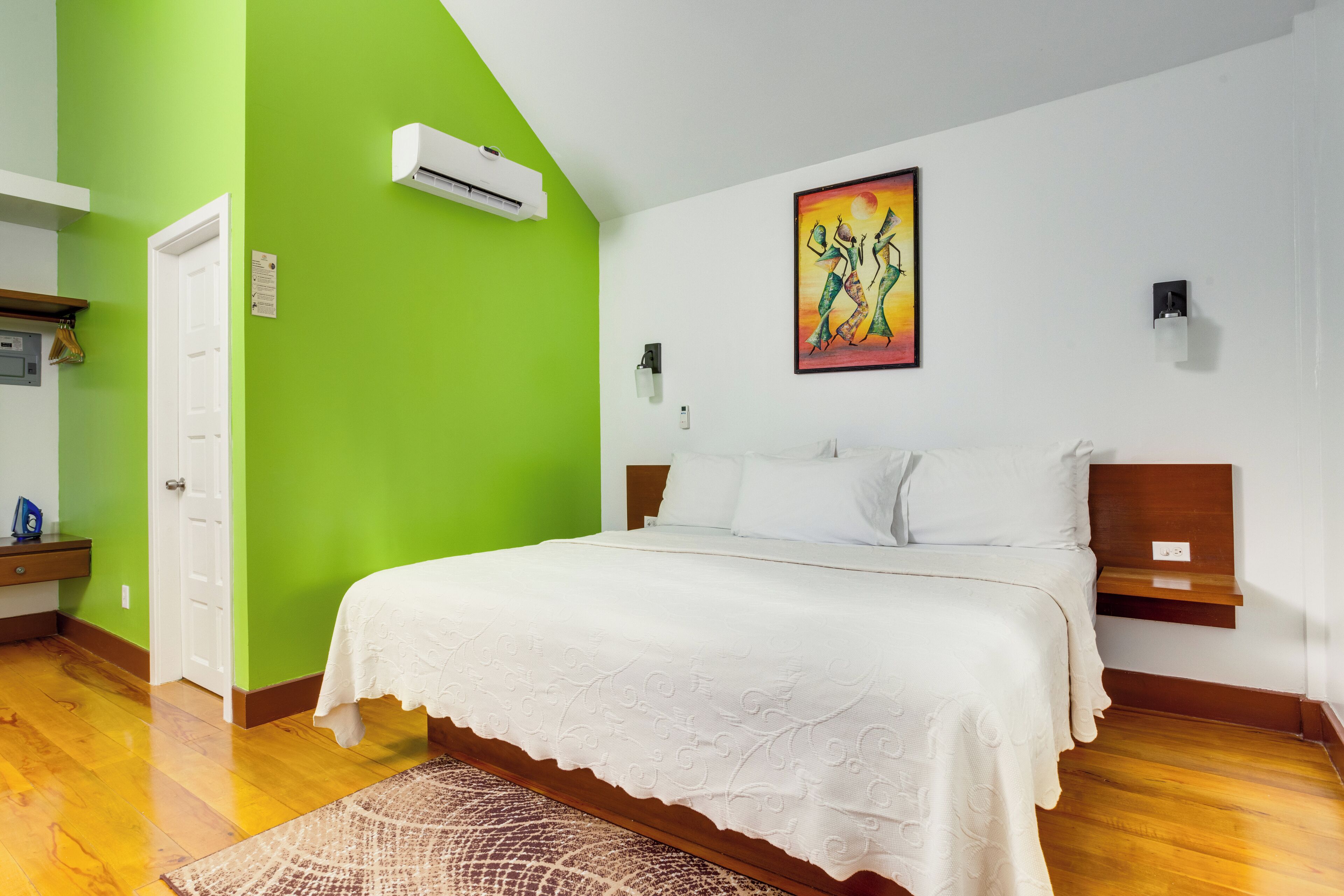 Un dormitorio con pared verde, cama blanca con cubrecama blanco, cabecera de madera y un cuadro sobre la cama.
