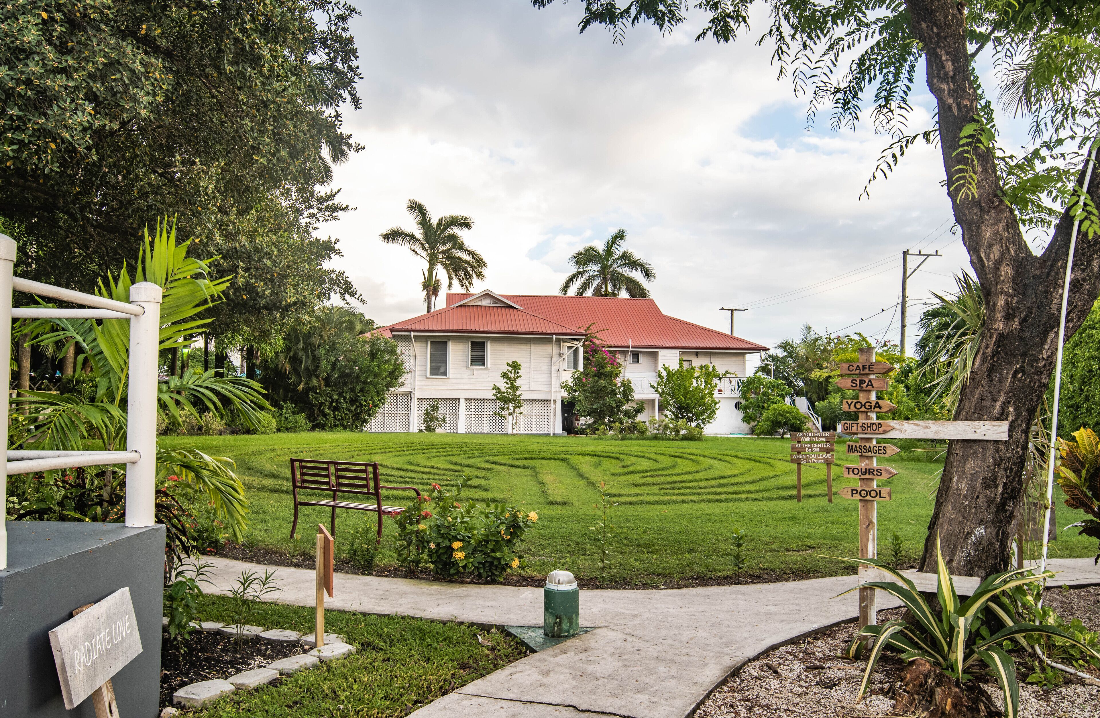 A white house with a red roof, surrounded by a well-maintained lawn and palm trees. A signpost lists amenities including a cafe, spa, yoga, gift shop, massages, tours, and pool.