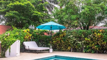 A swimming pool with a blue umbrella, a white lounge chair, and lush greenery in the background.