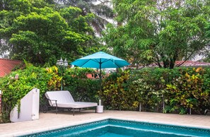 Una piscina con una sombrilla azul, una silla de descanso blanca y abundante vegetación de fondo.
