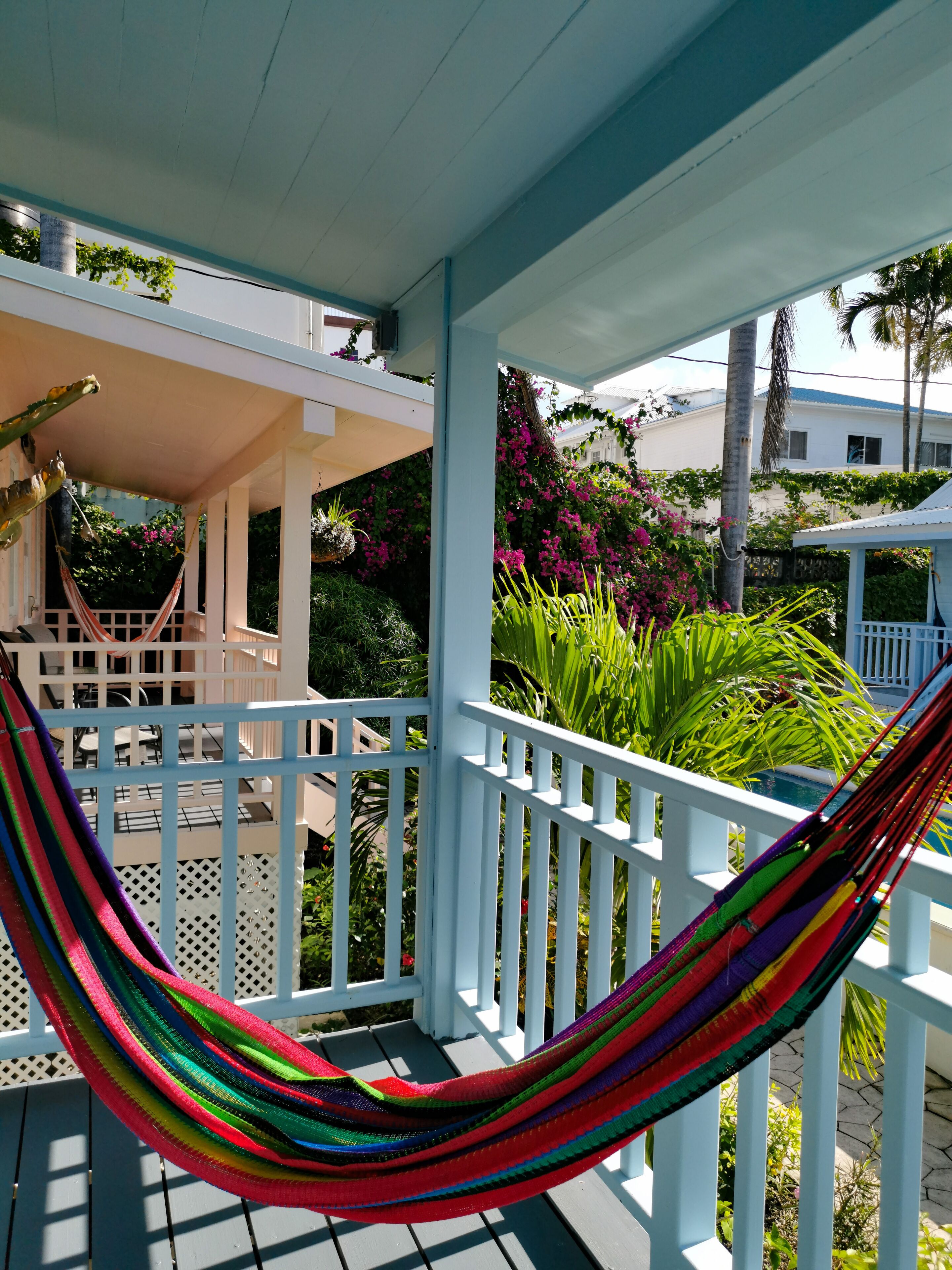 A hammock on a porch with a view of a garden and a building in the background.