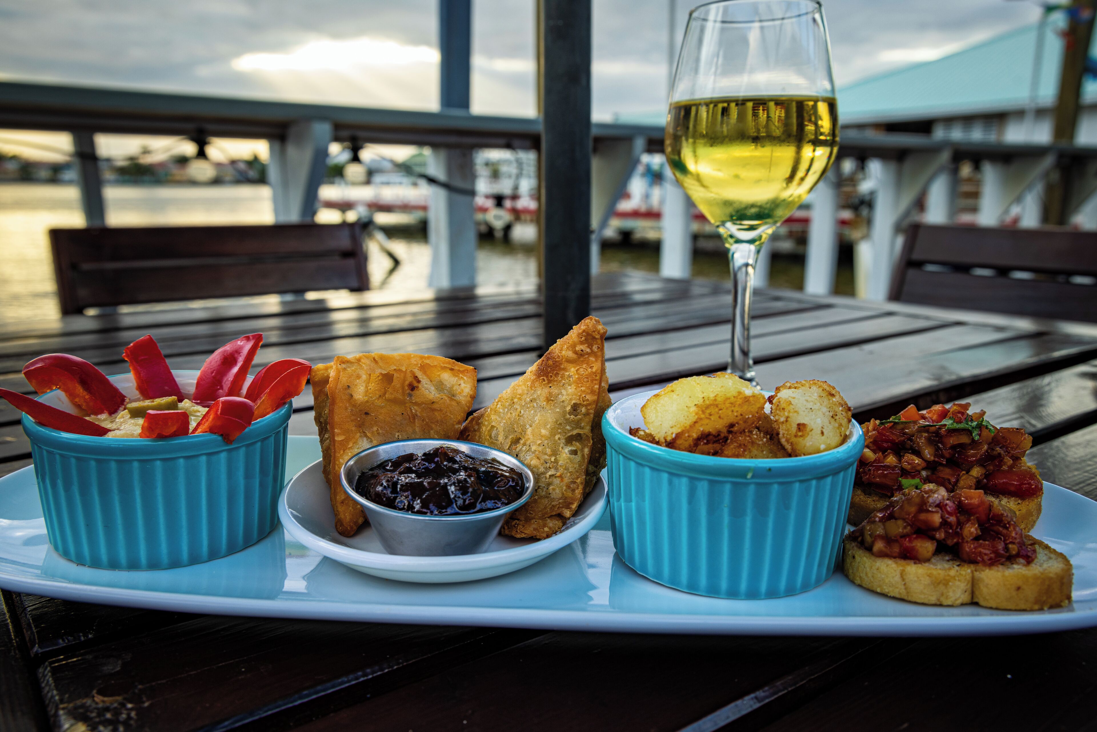 A meal with bread, dips, and a glass of white wine on a wooden table with a view of the water and boats.