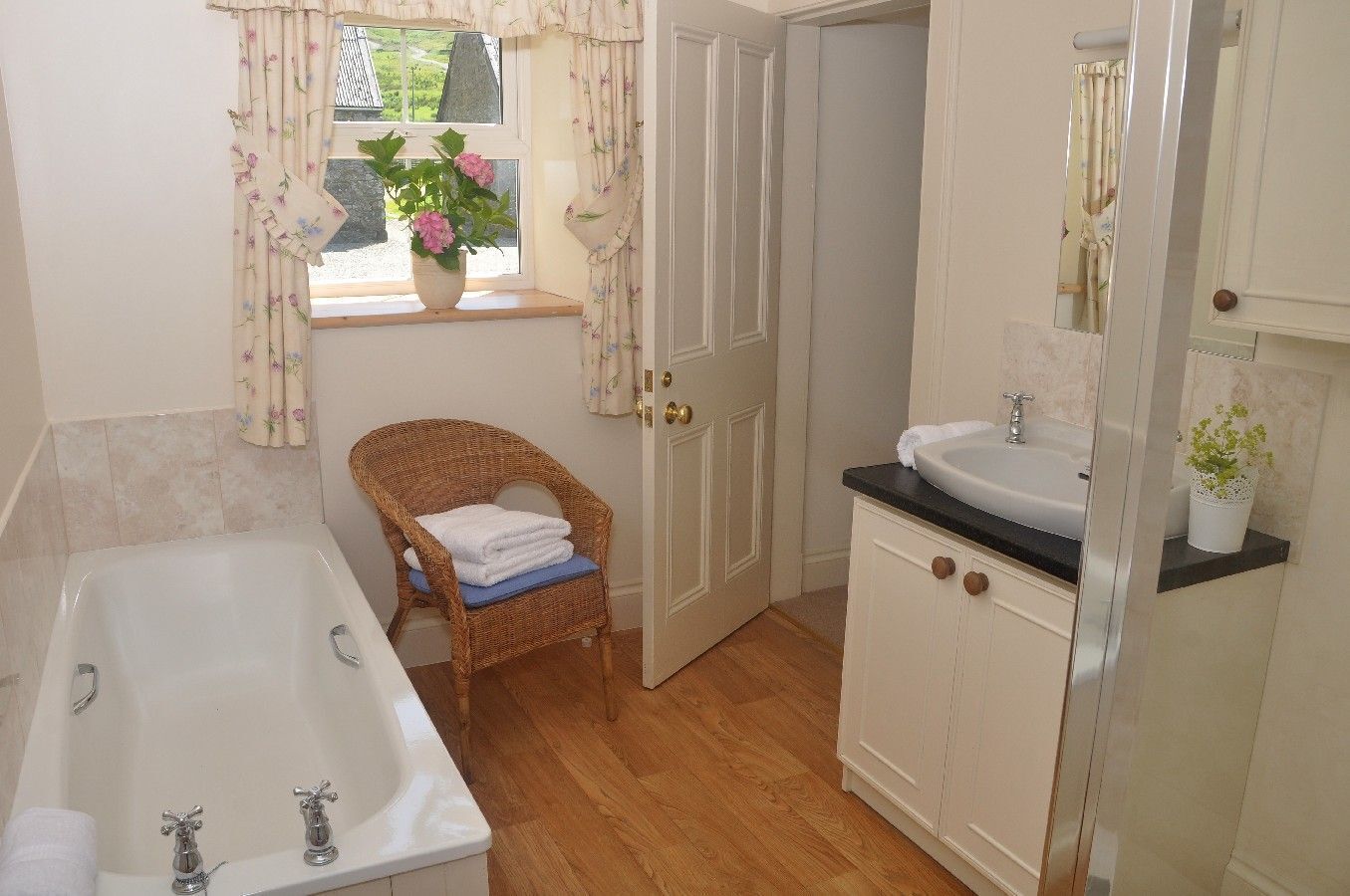 Ein Badezimmer mit einer Badewanne, einem Korbstuhl mit Handtüchern, einem Waschbecken und einem Fenster mit Vorhängen.