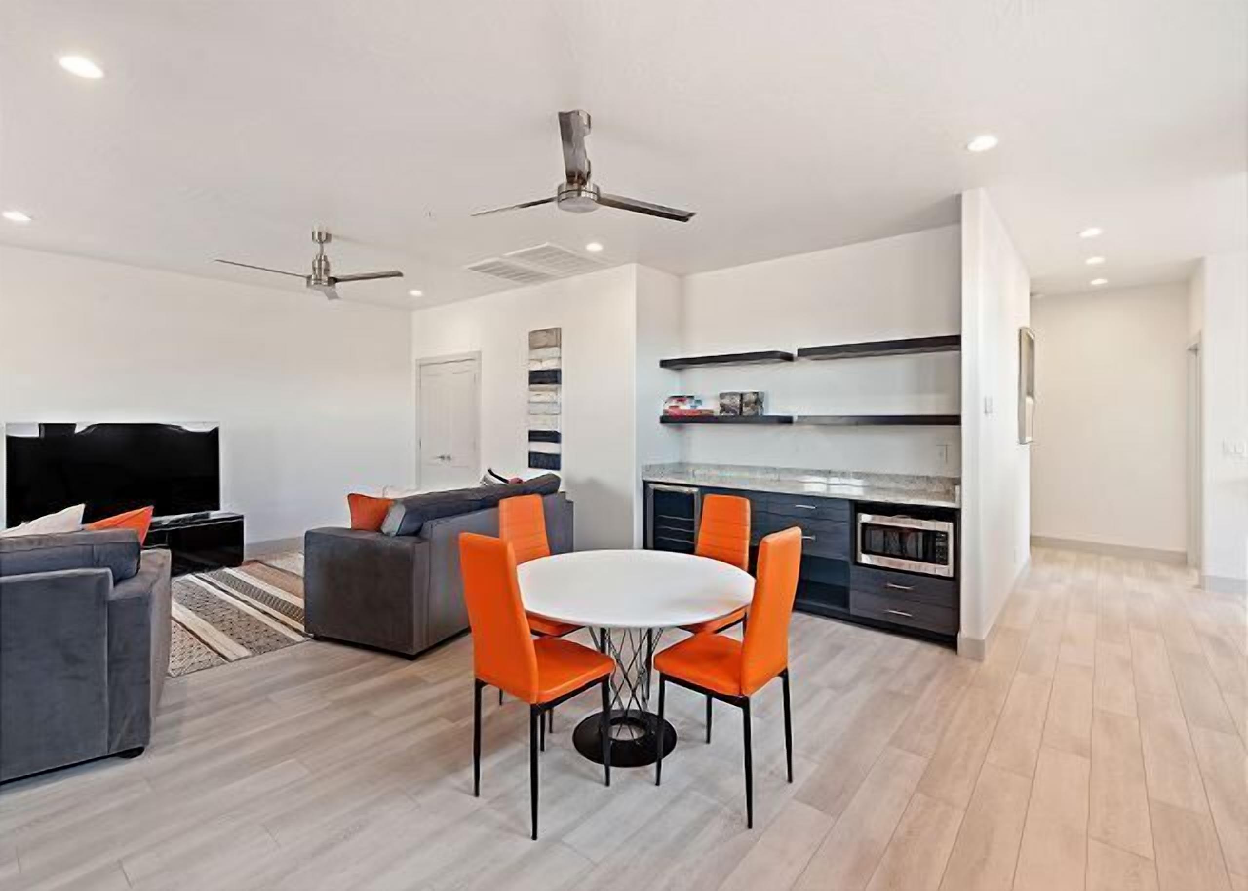 Una sala moderna con un sofá gris, sillas naranjas y un televisor. Hay ventiladores de techo, una cocina con armarios oscuros y una puerta blanca.