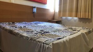 A hotel room with a large bed, a wooden headboard, a wall-mounted light, a window with curtains, and a colorful abstract painting above the bed. - Thermas Paradise (Rio Quente)