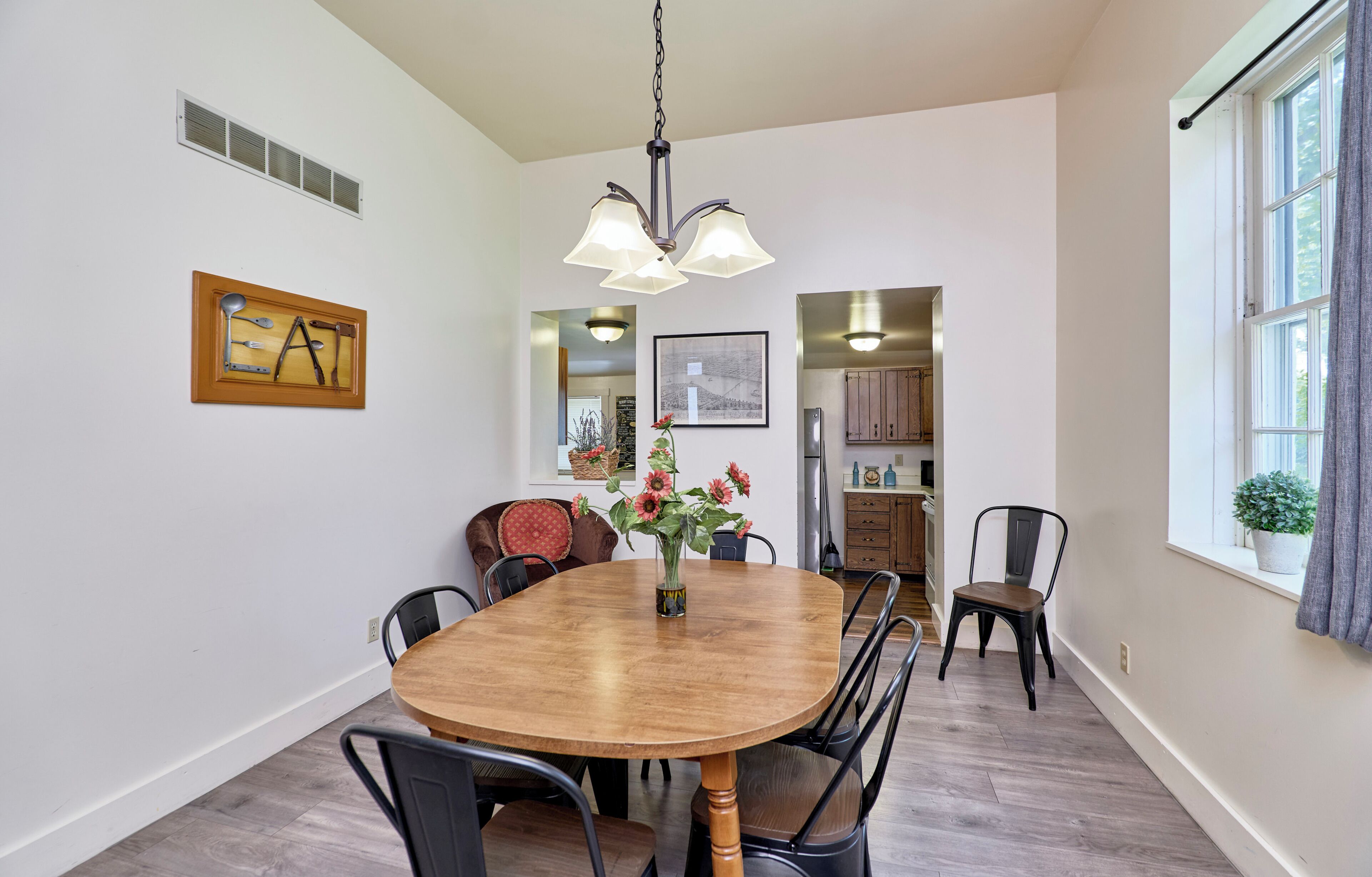 A dining area with a wooden table, chairs, a vase with flowers, a wall-mounted picture, and a window with curtains.