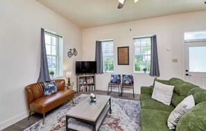 A living room with a brown leather sofa, a green sofa, a coffee table, and a TV. There are two windows with curtains, a bicycle decoration, and a bookshelf with books.