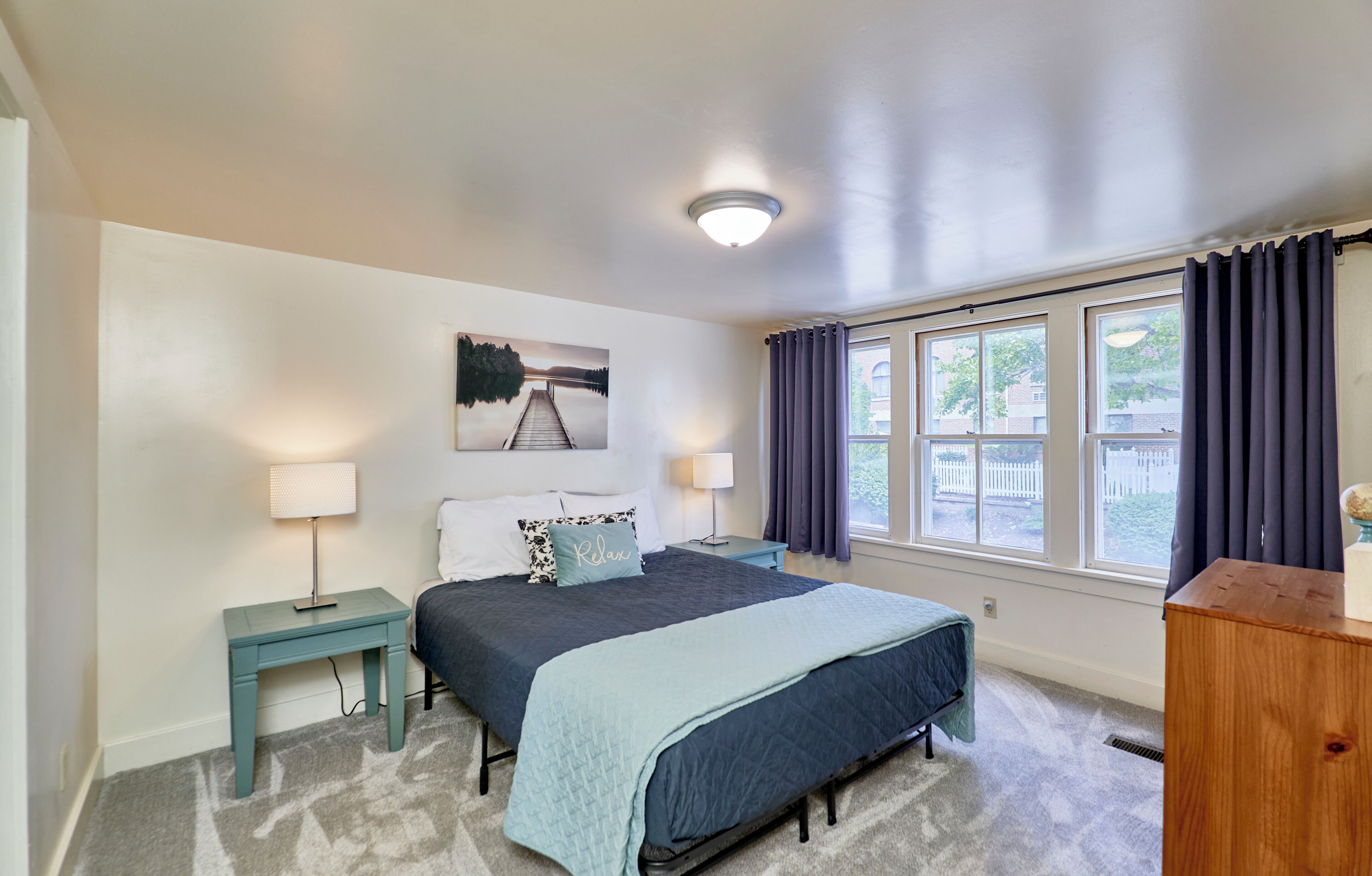 A bedroom with a bed, two bedside tables, a lamp, a painting, and a window with curtains.