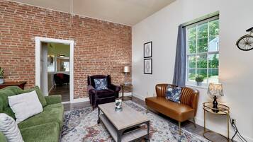 A living room with a brick wall, a green sofa, a brown leather sofa, a coffee table, and a window with a view of greenery.