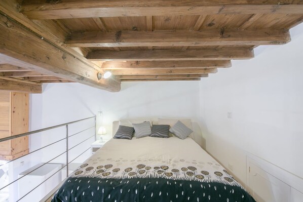 A bedroom with a wooden ceiling, a bed with patterned bedding, a nightstand, and a railing.