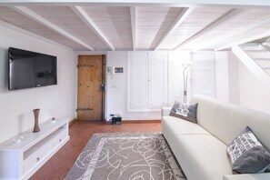 A living room with a flat-screen TV, a wooden door, white walls, a white sofa, and a patterned rug.