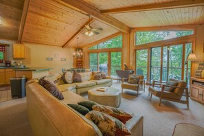 Ein gemütliches Wohnzimmer mit einer großen Couch, Hocker und Sesseln, Holzbalkendecke und großen Fenstern mit Blick ins Freie.