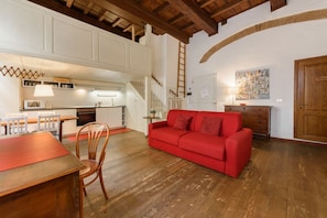 A spacious living area with a red sofa, wooden flooring, a dining table with chairs, a kitchen in the background, and a staircase.