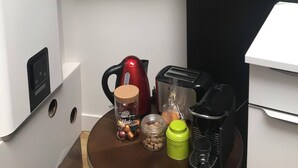 Ett köksutrymme med en kaffebryggare, vattenkokare, en burk med snacks och en termos på bänkskivan. Ovanför finns hyllor där glas och skålar förvaras.