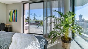 Une chambre d’hôtel moderne avec un grand lit, une plante en pot, un balcon offrant une vue sur la ville et une peinture abstraite aux couleurs vives accrochée au mur.