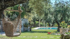 Eine Hängematte, die an einem Baum in einem Garten befestigt ist, mit Pool und Blumen im Hintergrund.