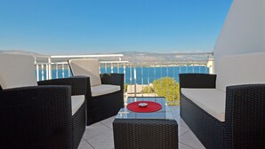 A balcony with wicker furniture and a glass table, overlooking a body of water and distant hills.