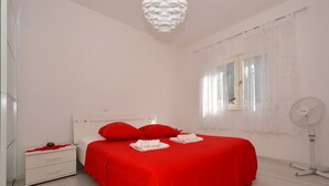 A room with a bed, a nightstand, a bedside table, a window with curtains, and a standing fan.