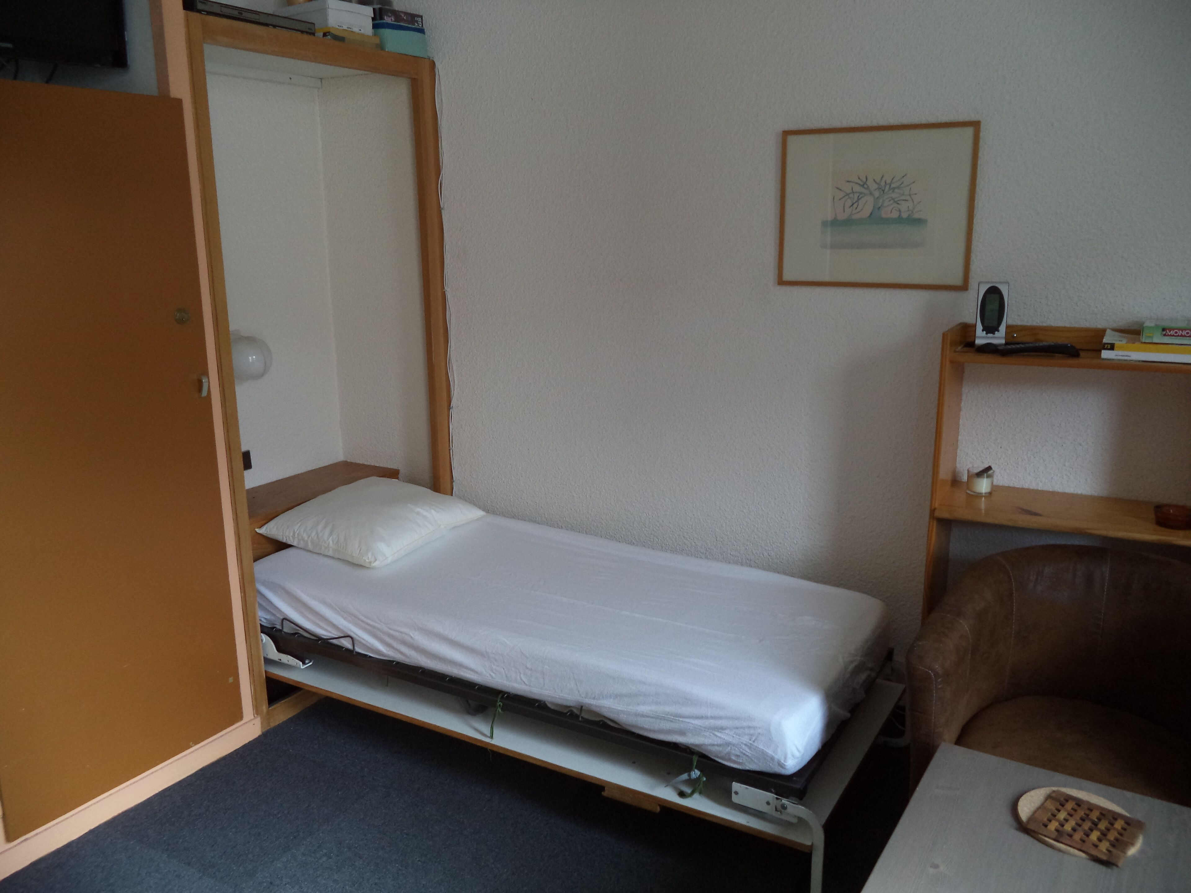 Una piccola camera con un letto singolo, una struttura letto in legno, una scrivania con sedia, uno scaffale con libri e una radio, e una porta che dà su un’altra stanza.