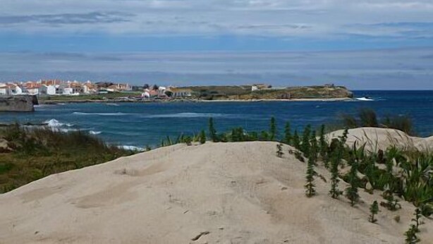 Eine Küstenlandschaft mit Sandstrand, einer kleinen Siedlung mit weißen Gebäuden und dem Meer im Hintergrund.