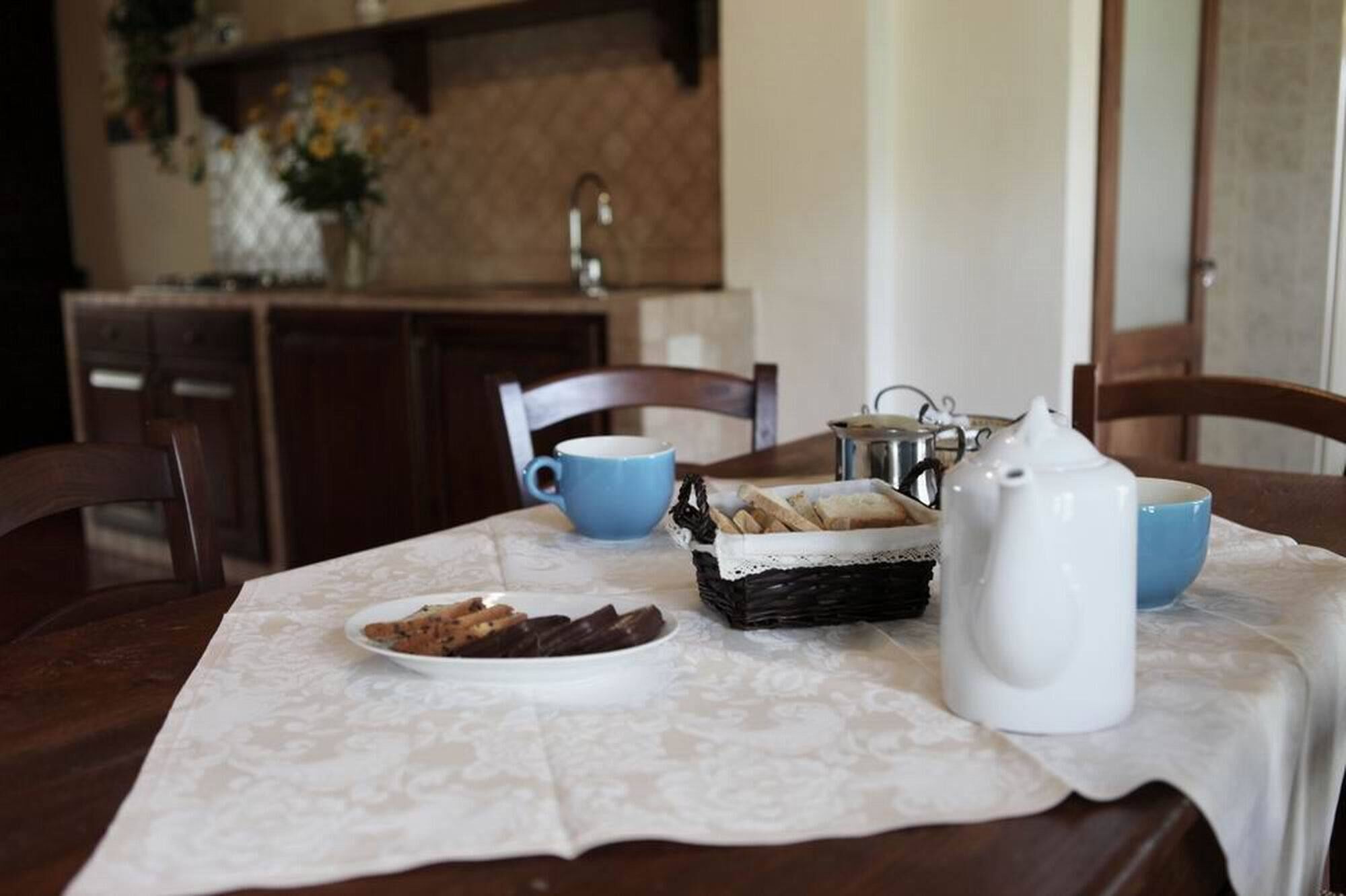 Une table à manger recouverte d’une nappe en dentelle, une assiette de biscuits, une tasse bleue, une théière blanche et un panier de pain.
