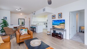 Ein modernes Wohnzimmer mit einem Ledersofa, einem Essbereich mit Korbstühlen, einer Küche mit weißen Schränken und einem großen Fernseher, der eine Strandansicht zeigt.