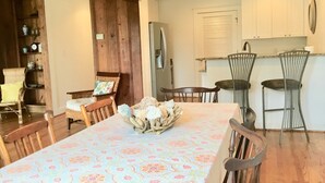 A dining area with a table set for four, surrounded by wooden chairs. A wicker chair with a cushion is placed at the far end. - Charming Beach Cottage Only 100 yds from the beach (Virginia Beach)
