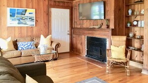 A wooden-paneled living room with a fireplace, a television mounted above it, and a sofa with patterned cushions. - Charming Beach Cottage Only 100 yds from the beach (Virginia Beach)