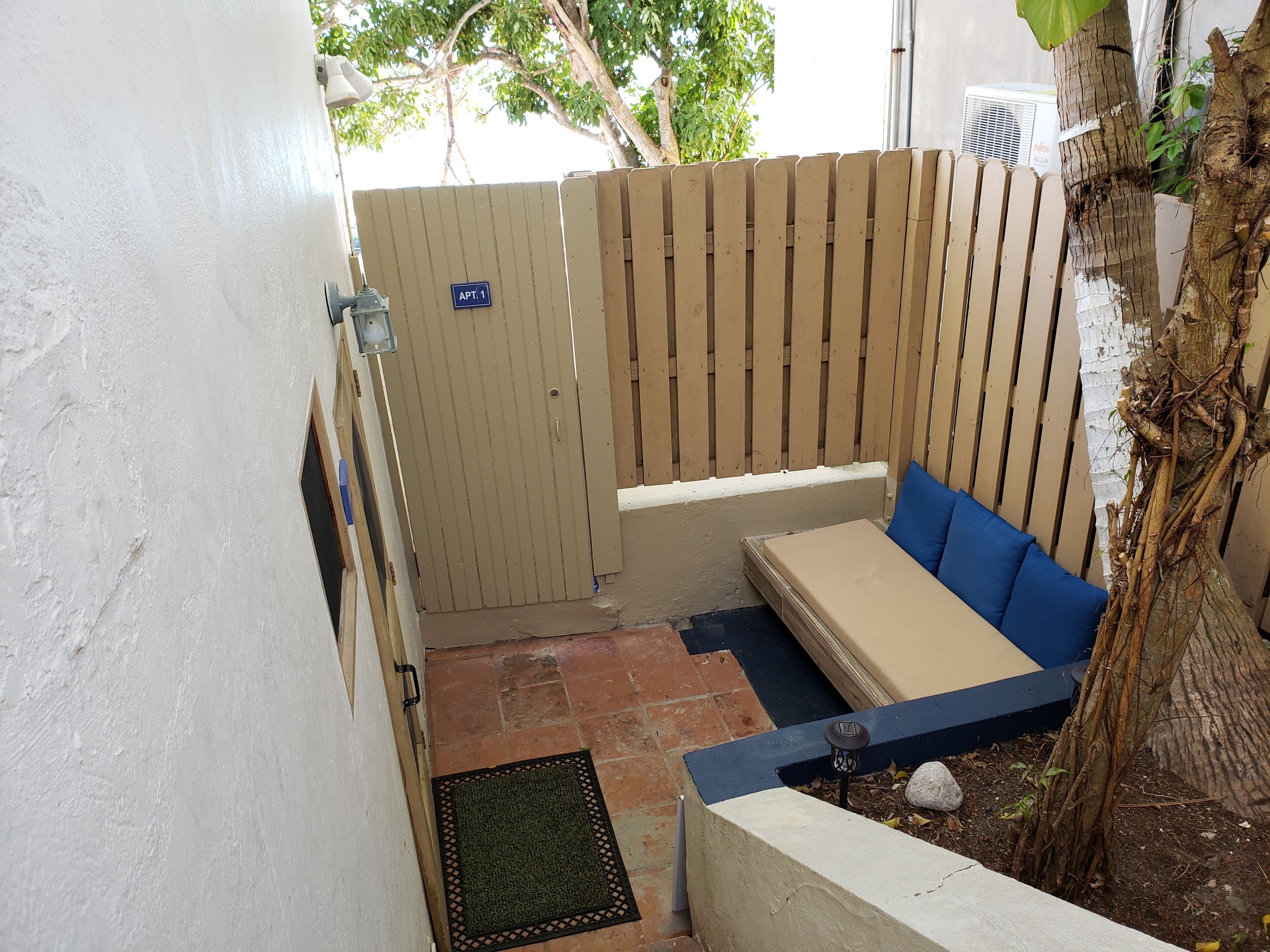 Un pequeño espacio exterior con una banca y una mesa cubierta, una valla de madera y una puerta con la inscripción 'APT. 1'.