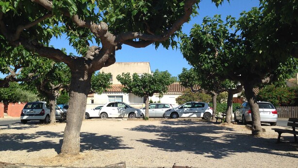 Un espace de stationnement avec plusieurs voitures, des arbres et un bâtiment doté d’un toit recouvert de tuiles.