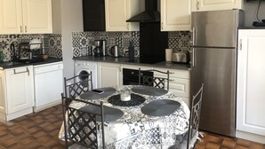 Une cuisine compacte avec des meubles blancs, un réfrigérateur en acier inoxydable, une machine à café et une table recouverte d’une nappe à motifs.