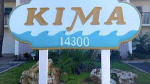 A sign with the name "KIMA" and the number "14300" in front of a building with balconies and palm trees.