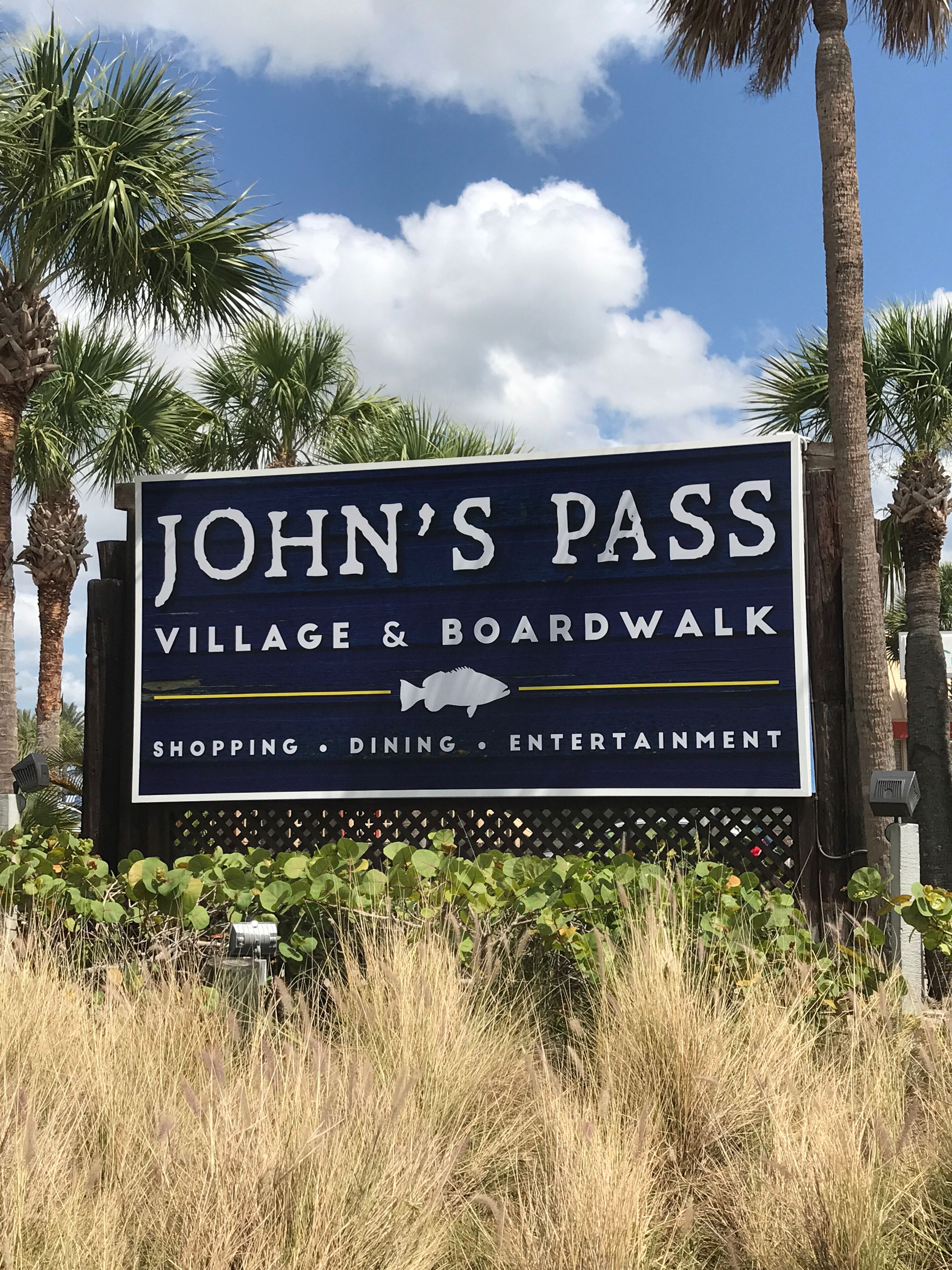 A sign for John's Pass Village & Boardwalk, highlighting shopping, dining, and entertainment.
