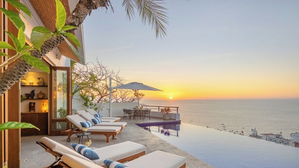 A seaside infinity pool with lounge chairs and a dining area overlooking the ocean at sunset.
