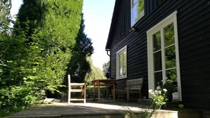 En träveranda med ett bord och stolar, omgiven av frodig grönska och ett mörkt trähus med vitmålade fönster.