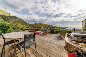 An outdoor patio with a wooden table and chairs, a barbecue grill, and a view of a hillside and water.