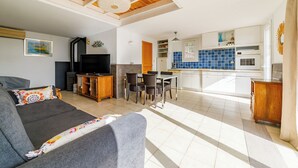 Un grand espace de vie comprenant un canapé, une télévision et une table à manger. On aperçoit une cuisine aux carreaux bleus et un balcon à l’arrière-plan.