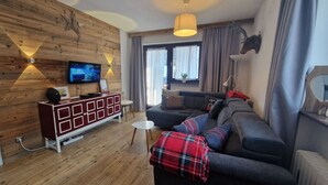 Un salon moderne avec un mur en bois, un téléviseur fixé au-dessus d’un meuble rouge et blanc, un canapé gris avec une couverture à carreaux et une grande fenêtre avec des rideaux.