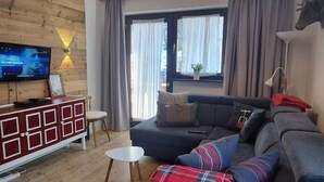 Un salon moderne avec un mur en bois, un canapé gris, un plaid à carreaux rouges et bleus, une petite table blanche et un téléviseur fixé au mur.