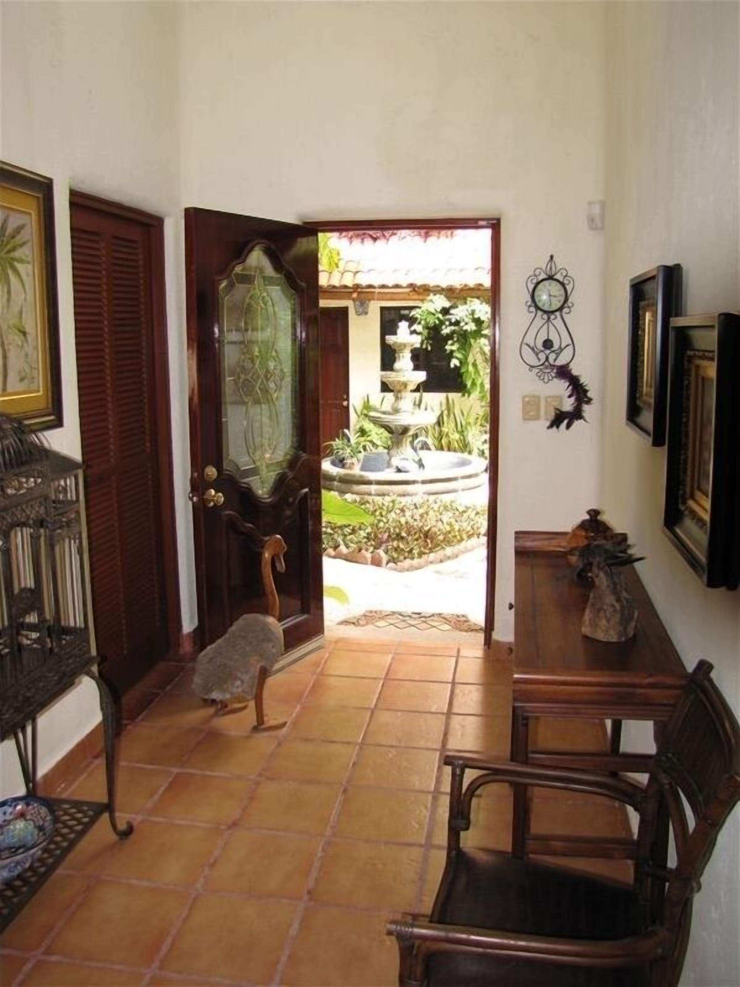 A room with a wooden door, a small table, and a chair. There is a tiled floor and a window with a view of a garden with a fountain.