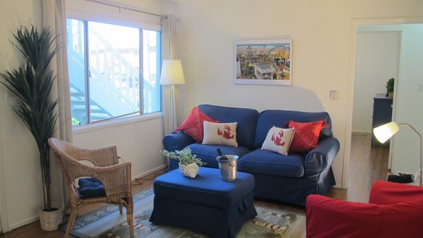 Una sala de estar con un sofá azul, cojines rojos, un sillón de mimbre, una mesita y un ventanal con cortinas.
