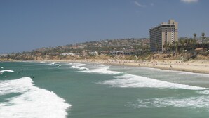 Escena de playa costera con olas, una orilla arenosa y un edificio al fondo.