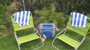 Dos sillas de jardín verdes con toallas a cuadros azules y blancas, una mochila gris y una botella de agua sobre una zona cubierta de césped con plantas al fondo.
