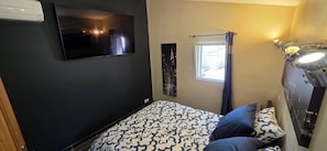 Ein Schlafzimmer mit Bett, einem an der Wand montierten Fernseher, einem Fenster mit Vorhängen und einer Deckenleuchte.