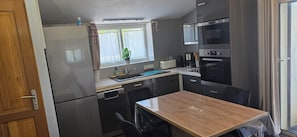 Eine kleine Küche mit Kühlschrank, Herd, Mikrowelle und Spüle. Ein Fenster mit einer Pflanze, ein Tisch mit Stühlen und eine Tür sind ebenfalls vorhanden.