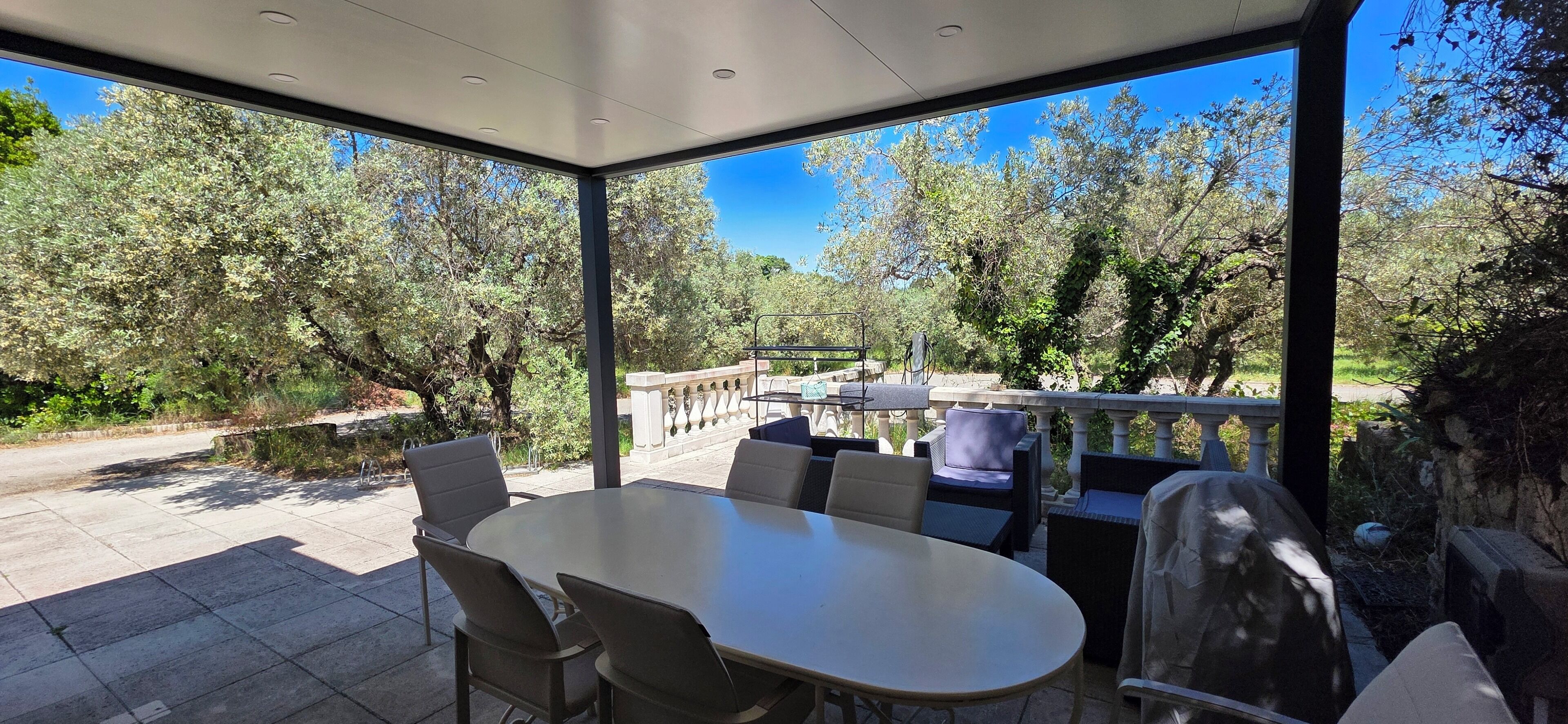 Un espace extérieur abrité, aménagé pour les repas, avec une table et des chaises, donnant sur un jardin avec des arbres et un sentier en pierre.