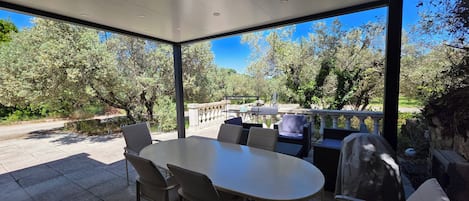 A covered outdoor dining area with a table and chairs, overlooking a garden with trees and a stone pathway.